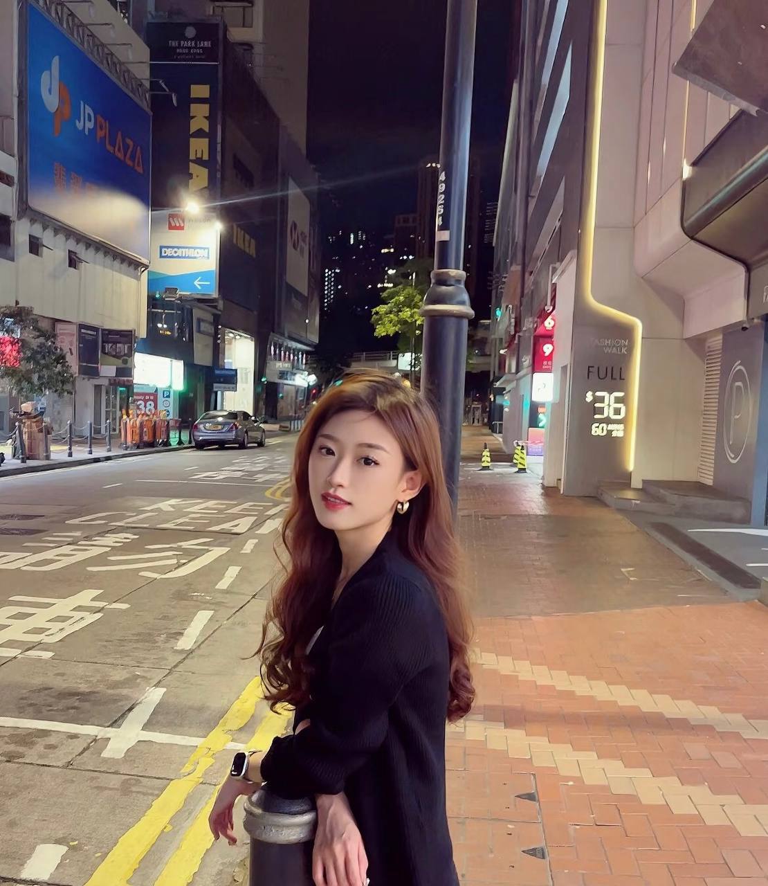 Portrait in a black knit cardigan, leaning against a bollard on a city street at night