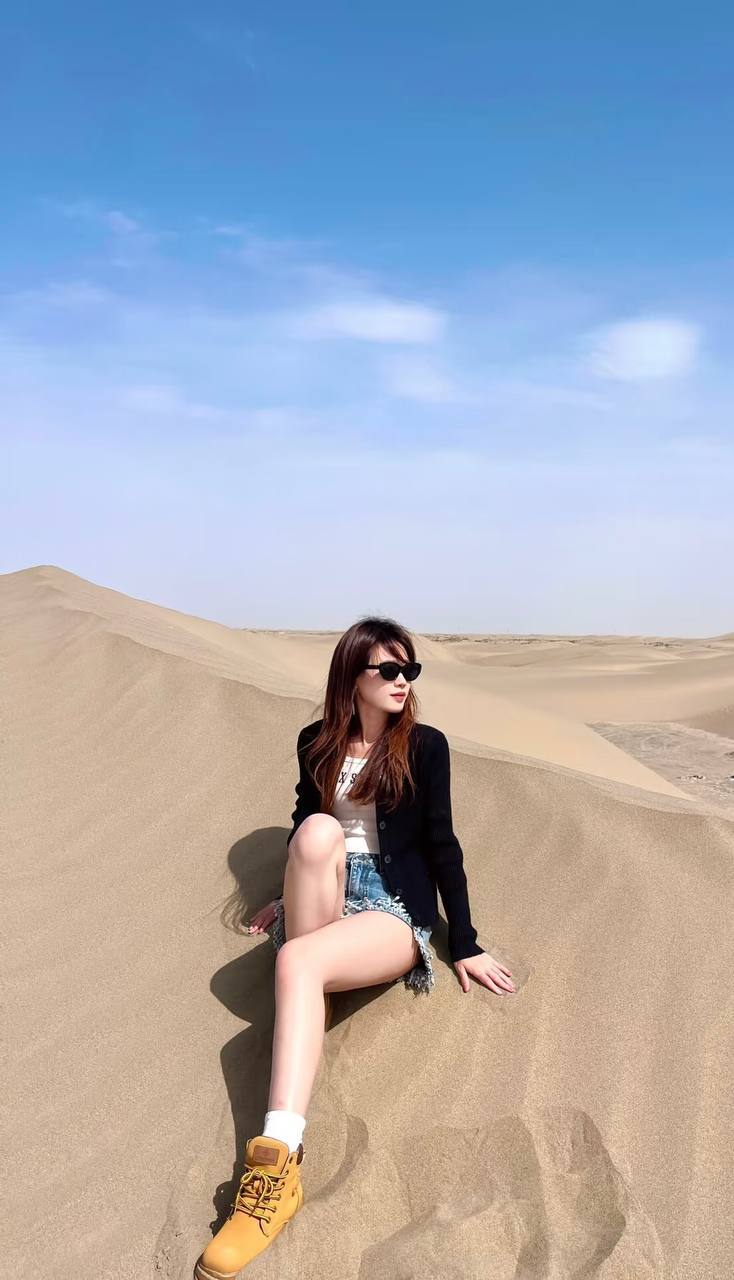 Full-length travel photo sitting on a sand dune, wearing sunglasses