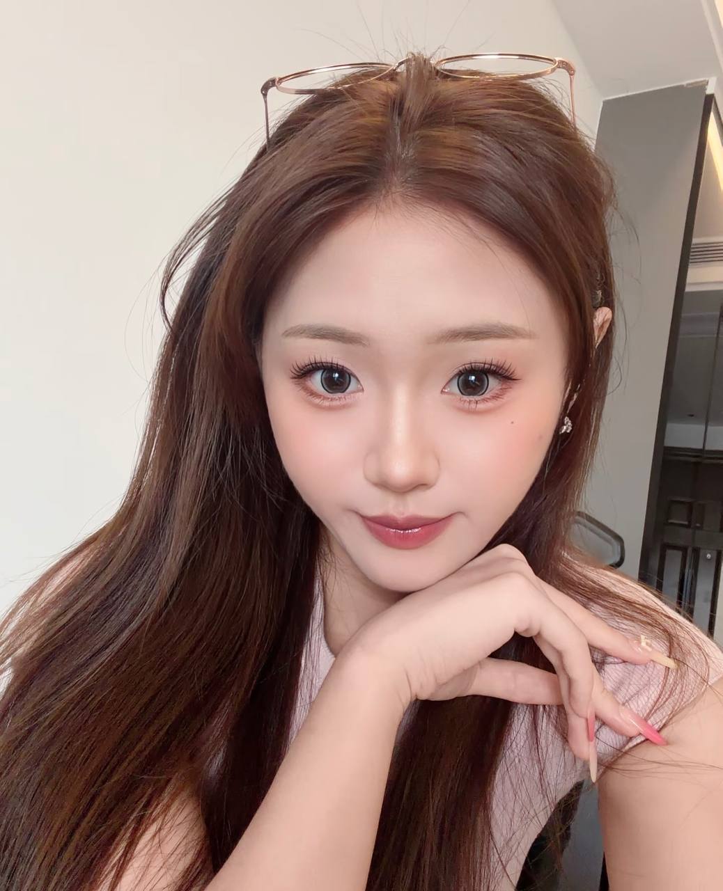 Selfie in a pale pink top, with glasses resting on the head and chin resting on a hand
