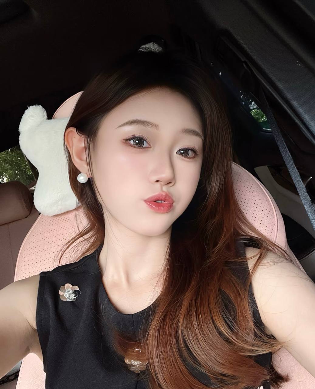 Selfie in a black sleeveless top and pearl earrings, with a pink car seat in the background