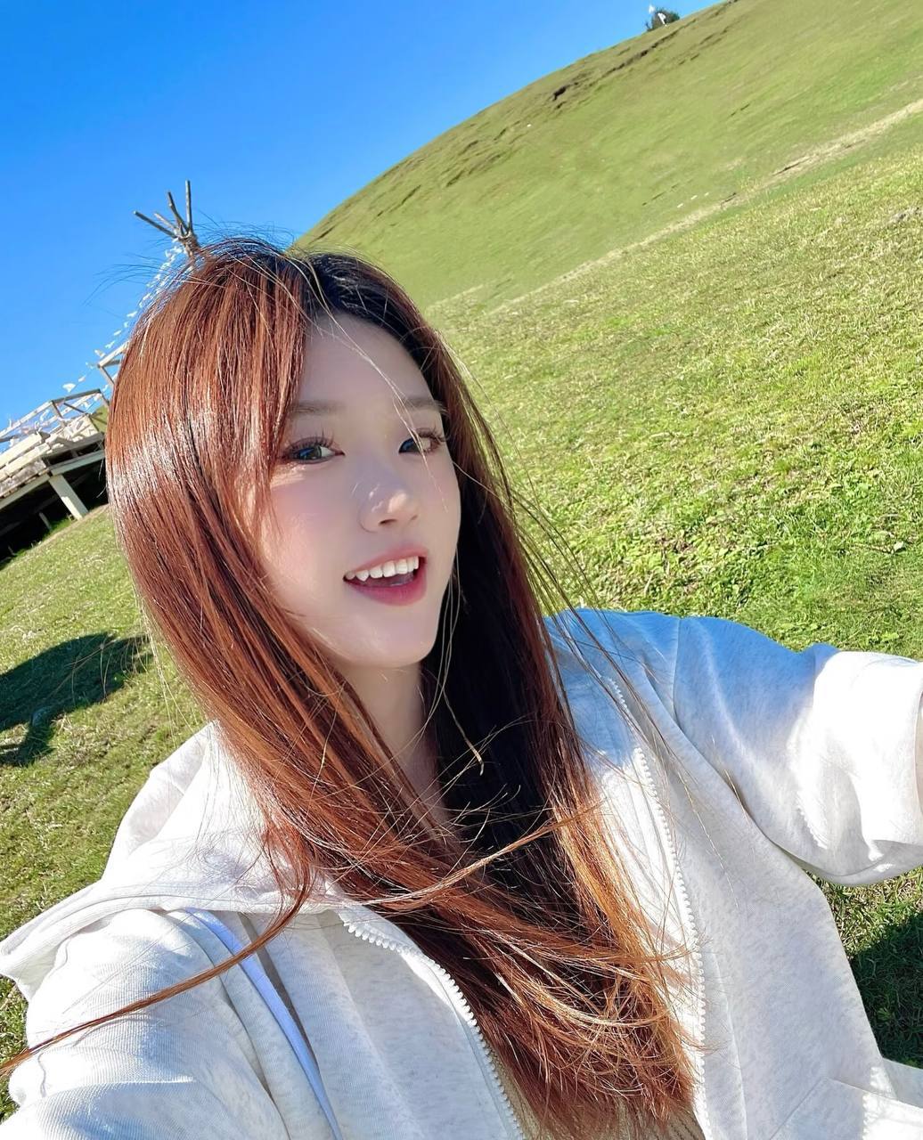 Smiling selfie in a light-colored hoodie, with green hills and blue skies