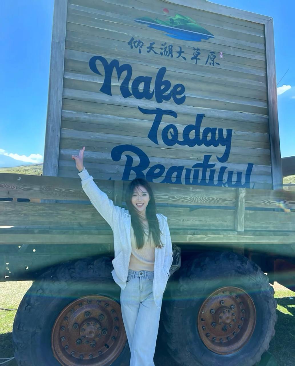 Full-length photo smiling with one arm raised in front of a wooden sign reading Make Today Beautiful